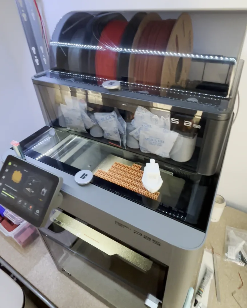 La Bambu Lab P2S nueva de Erwan en la estantería del taller, con el AMS 2 Pro encima y una tanda de piezas imprimiéndose
