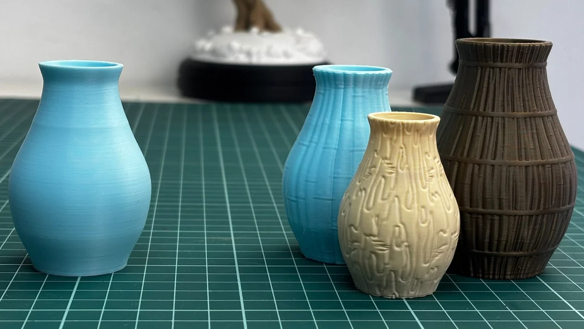 Cuatro jarrones impresos en 3D sobre una alfombrilla de corte: uno azul liso, uno azul con estrías, uno crema con textura de Corteza y uno marrón