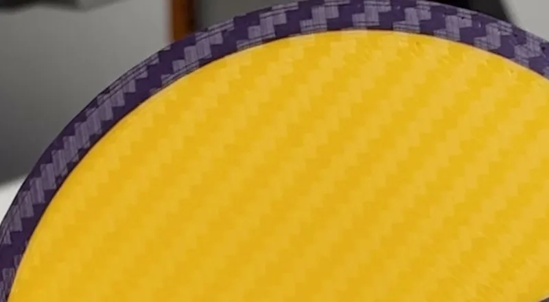 Primer plano de la cara de abajo de una pieza naranja impresa en 3D, con el dibujo de fibra de carbono copiado de la placa y el borde morado con el mismo dibujo