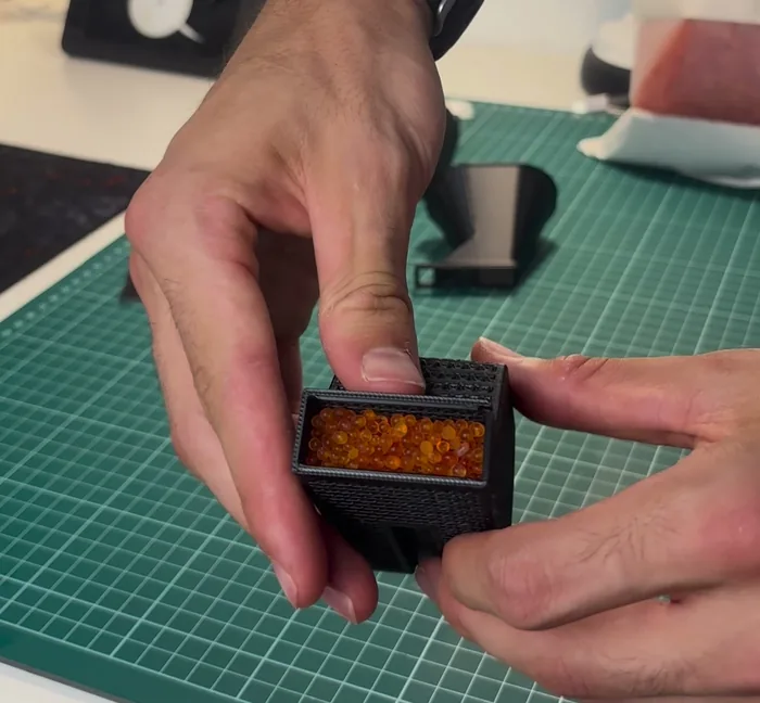 Depósito negro impreso en 3D lleno de perlas naranjas de gel de sílice, sujeto con las manos