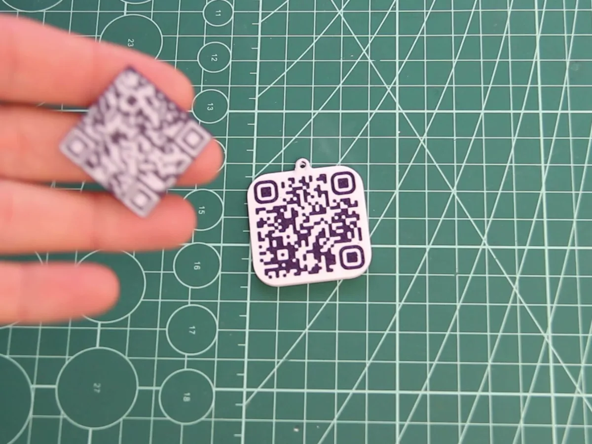 Dos llaveros con código QR impresos en blanco y negro, uno en la mano y otro sobre la alfombrilla de corte